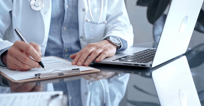 Male doctor wearing white medical coat over a blue shirt is writing medical records on a glass desk with laptop.