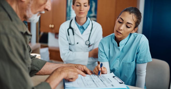 Young nurse assisting senior man to fill out medical documents at reception desk at doctor's office.